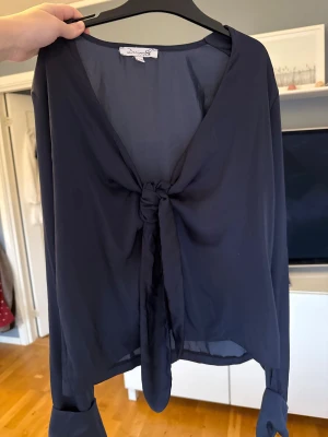 Mörkblå omlottblus från Design by Si - Säljer en stilren mörkblå blus från Design by Si i storlek XS/S. Blusen har omlottmodell med snygg knytning framtill och långa ärmar. Materialet är mjukt och har en lätt glansig finish som ger en lyxig känsla. Perfekt för dig som gillar trendiga detaljer och enkel design.
