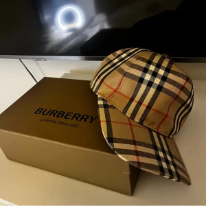 Burberry keps - Snygg Burberry keps i klassiskt rutigt mönster med beige, svart, röd och vit färg. Kepsen har böjd skärm och justerbar rem bak. Tillverkad i bomull med ikonisk Burberry-design som ger en lyxig vibe till din outfit.