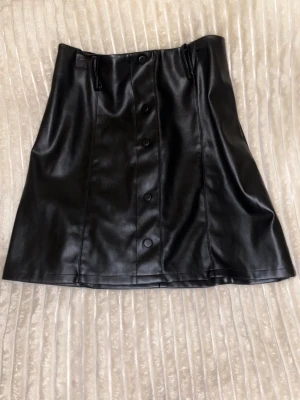 Svart kort skinn kjol 🖤🤩 - Snygg svart kort kjol i skinnimitation. Kjolen har tjock material, dekoration knappar framtill, bälteshällor och en väldigt fin skräddarsydd kedja. Detta är THE Stockholm girl style! En edgy eller en old money look du väljer hur du ska styla den! Kjolen är väldigt bekväm och materialet är mjukt inuti. Använd endast 1 gång så den är i princip i ny skick! Fick så många komplimanger på eventet.⭐️😍  Längd 42cm, Bredd från mitten på kjolen ~47cm, Midja 35cm
