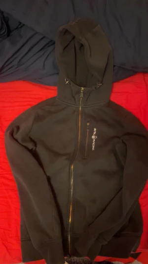 Svart hoodie från Sail Racing, S - Svart hoodie från Sail Racing i storlek S. Tröjan har hel dragkedja framtill, bröstficka med dragkedja och broderad logga. Utrustad med huva och långa ärmar. Perfekt för kyliga dagar och snygg till jeans. Den är i väldigt bra skick