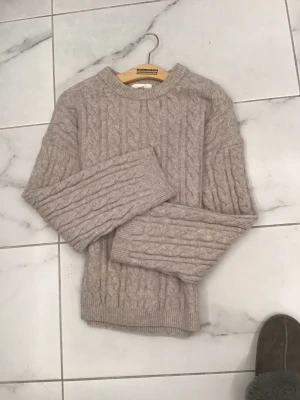 Beige kabelstickad tröja H&M XS - Snygg beige kabelstickad tröja från H&M i storlek XS. Tröjan har rund hals, ribbade muddar och ett klassiskt flätmönster över hela plagget. Perfekt för dig som gillar en chill och stilren look under höst och vinter.