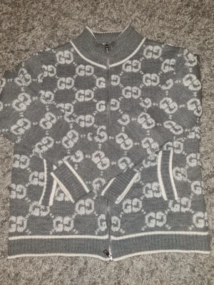 Grå kofta med GG-mönster och dragkedja - Snygg grå kofta från Gucci med vit GG-logga över hela tröjan. Koftan har ribbade muddar, ståkrage och hel dragkedja framtill. Fina vita detaljer vid fickor, krage och nederkant. Perfekt för dig som gillar lyxiga och trendiga plagg.