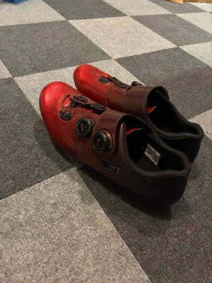 Röda Shimano cykelskor med BOA - Shimano cykelskor i metallicrött med svarta detaljer och dubbla BOA-spännen för smidig justering. Ovandelen är perforerad för ventilation och insidan är vadderad för extra komfort. Snygg, sportig design som passar dig som gillar att cykla med stil.