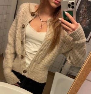 Beige stickad kofta från Zara - Supermysig beige stickad kofta från Zara med stora bruna knappar och ribbade muddar. Koftan har en croppad passform och lång ärm, perfekt att slänga över en topp när det är kyligt. Enkel att styla och riktigt skön att ha på sig.