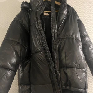 Svart vinterjacka med bälte - Säljer en svart, glansig pufferjacka från Nelly med huva och bälte i midjan. Jackan har långa ärmar, dragkedja framtill och en oversized passform. Helt ny men utan prislapp. Säljs då lappen råkades klippas bort för tidigt så retur ej möjlig. 