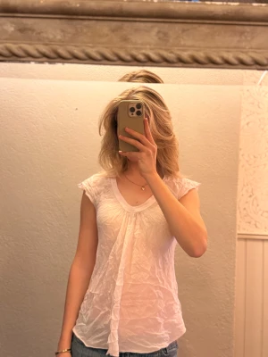 Vit blus från Nolita med volang - Säljer en söt vit blus från Nolita i 100% bomull. Blusen har korta ärmar, v-ringad hals och en snygg volangdetalj framtill. Tunt och luftigt tyg med lätt skrynklig look, perfekt för dig som gillar romantisk och feminin stil.