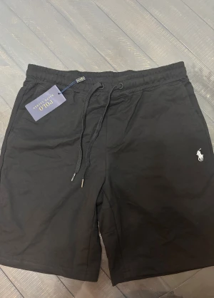 Svarta Polo Ralph Lauren Shorts - Svarta shorts från Polo Ralph Lauren med klassisk logga broderad i vitt på vänster ben. Modellen har snörning i midjan, elastisk linning och en bakficka. Tillverkade i mjuk bomull för skön känsla och avslappnad passform. Perfekta för chill dagar eller träning.