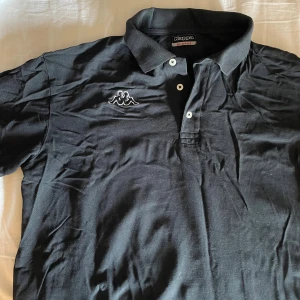 Svart pikétröja från Kappa - Svart pikétröja från Kappa i storlek XL med klassisk krage och två vita knappar framtill. Tröjan har Kappas ikoniska broderade logga på bröstet och är gjord i mjuk bomull. 