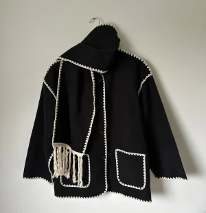 Svart kappa med vit kant och halsduk - Unik svart kappa med halsduk med dekorativa vita kanter och två stora fickor fram. Medföljer matchande sjal med fransar. Rak passform och rund halsringning, stängs med knappar. Perfekt för dig som gillar statement-plagg med retrovibbar. 5K7