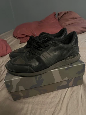 Valentino Rockrunner svarta sneakers - Säljer ett par svarta Valentino Rockrunner sneakers med coola nitar på hälen och kamouflagemönster på ovansidan. Skorna har snörning och är tillverkade i skinn och mocka, med platt sula och rund tå. Box med camo-print och dustbag ingår.