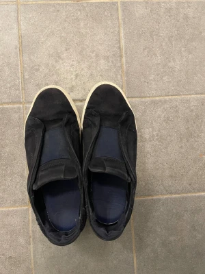 Svarta sneakers från Axel Arigato - Svarta slip-on sneakers från Axel Arigato med vit platt sula och rund tå. Ovandelen är i mocka och textil, med diskret logga på sidan. Snygg minimalistisk design som passar till jeans eller shorts. Bekväma och stilrena för dig som gillar cleana sneakers.