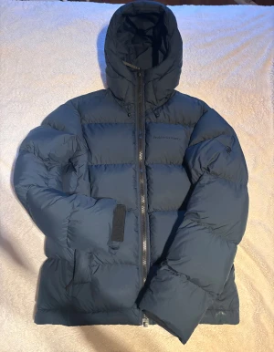 Mörkblå Peak Performance dunjacka - Säljer en mörkblå dunjacka från Peak Performance med tecken på användning, alltså att tyget har blivit blankt längst nedre delen av ärmarna och midjan/fickorna.                                      Blixtlåslöparen är lite sliten som du ser på bilderna och det finns några små osynliga repor. Annars så fungerar allt som det ska och överlag så ser den fräsch ut. 