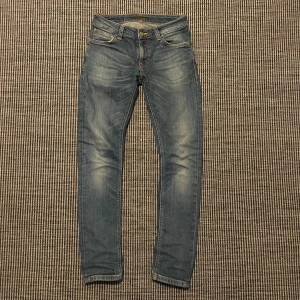 Blå  jeans från Nudie Jeans - Snygga blå jeans från Nudie Jeans med bra passform och klassisk femficksdesign. Jeansen har ljusa slitningar framtill och bak, samt Nudie Jeans logga på bakfickan och läderpatch i midjan. Perfekt för dig som gillar en smal siluett och stilren look. Storlek W27 L30