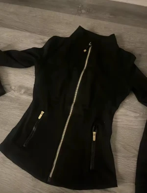 Svart slim vindjacka med gulddragkedja - Snygg svart vindjacka med guldfärgade dragkedjor framtill och på fickorna. Jackan har en slim passform och långa ärmar, perfekt för dig som gillar stilrena detaljer och sportig look. Passar dig som vill ha en jacka med lite edge och coola detaljer.