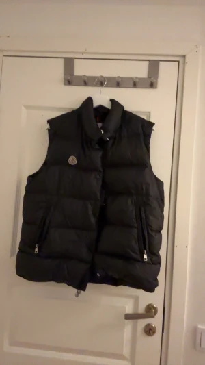 Svart dunväst från Moncler - Svart dunväst från Moncler med hög krage och quiltad design. Västen har två fickor med dragkedja, tryckknappar framtill och Moncler-logga på bröstet. Materialet är glansig polyester och dunfyllning, perfekt för kyliga dagar.