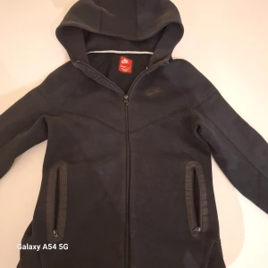 Svart hoodiejacka från Nike - Svart hoodiejacka från Nike med hel dragkedja framtill och två fickor med dragkedja. Jackan har en sportig look, huva och diskret Nike-logga på bröstet. Tillverkad i mjukt bomullsmaterial, perfekt för chill eller träning.