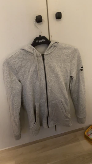 Grå hoodie från Adidas med dragkedja - Säljer en grå hoodie från Adidas med svart dragkedja och snörning i huvan. Tröjan har långa ärmar, två fickor framtill och Adidas-logga på vänster ärm. Perfekt för chill eller träning, mjukt material och klassisk look.
