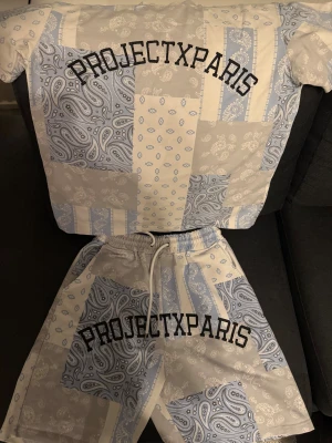 Blå och vita paisley shorts Project X Paris - Säljer ett par fräscha shorts från Project X Paris med patchwork paisley-mönster i ljusblått och vitt. De har elastisk midja med snörning och stor logga framtill. Perfekt för dig som gillar streetwear och unik design. Köpt i Spanien för 900kr
