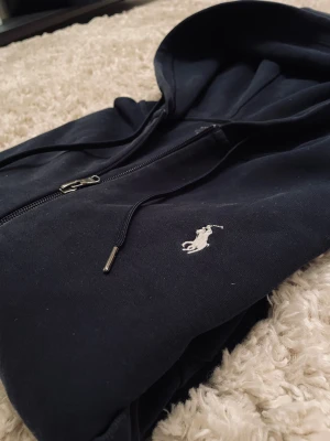Marinblå hoodie från Polo Ralph Lauren - Snygg marinblå hoodie från Polo Ralph Lauren med klassisk vit logobrodering på bröstet. Tröjan har huva med snörning, dragkedja framtill och två fickor. Tillverkad i mjuk bomull, perfekt för chill dagar. Långärmad och med ribbade muddar för extra komfort.