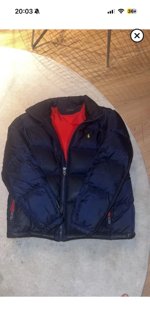 Mörkblå pufferjacka från Polo Ralph Lauren - Säljer en mörkblå pufferjacka från Polo Ralph Lauren med gul broderad logga på bröstet och röd insida. Perfekt för vintern. 