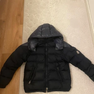 Mörkblå Moncler dunjacka med huva - Säljer en mörkblå dunjacka från Moncler med avtagbar huva och snygga detaljer som logotyp på ärmen och serietidningsetikett på insidan. Jackan har två fickor med dragkedja och quiltad design. Perfekt för kalla dagar och riktigt stilren.