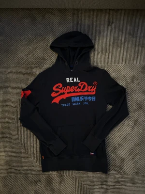 Svart Superdry hoodie  - Svart hoodie från Superdry med stor röd och vit logga samt japanska tecken på bröstet. Har huva, känguruficka och röd detalj på ena ärmen. Mjuk insida och klassisk passform, perfekt för chill dagar.