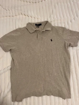 Beige pikétröja Polo Ralph Lauren L - Klassisk beige pikétröja från Polo Ralph Lauren i storlek L. Tröjan har korta ärmar, krage och knäppning framtill med två knappar. Broderad logga på bröstet. Skön bomullskvalitet och stilren passform.