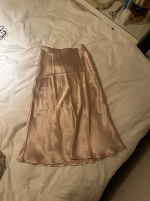 Champagnefärgad satinkjol - Säljer en glansig, champagnefärgad långkjol i satin med mjuk, följsam passform. Kjolen har en stilren och enkel design utan synliga detaljer, perfekt för dig som gillar en clean look. Den har lätt utsvängd form och vacker lyster.