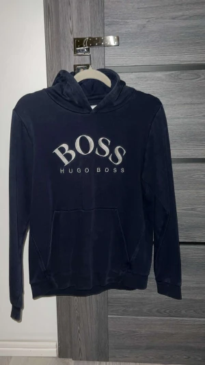 Mörkblå hoodie från Hugo Boss - Snygg mörkblå hoodie från Hugo Boss med stort BOSS-tryck i vitt på bröstet. Tröjan har huva, långärmade ärmar och en klassisk känguruficka framtill. Perfekt för dig som gillar stilrena märkesplagg och vill ha en chill look.