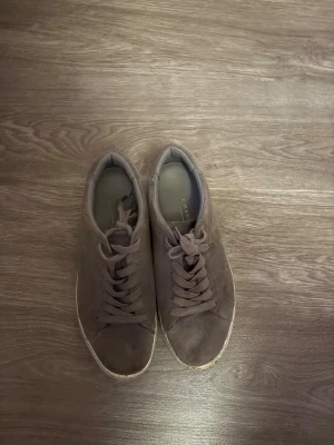 Bruna mockasneakers med snörning - Snygga bruna sneakers i mocka med platt sula och rund tå. Skorna har klassisk snörning och en vit kant runt sulan som ger en stilren look. Perfekta för dig som gillar enkel och clean stil. Märket F MAR syns på innersulan.