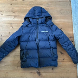 Marinblå pufferjacka Calvin Klein Jeans - Snygg marinblå pufferjacka från Calvin Klein Jeans med huva och logga på bröstet och ärmen. Jackan har dragkedja framtill, två fickor med dragkedja och quiltad design. Perfekt för kalla dagar och har en modern, clean look. Finns några små hål som knappt syns 