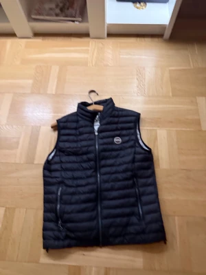 Svart quiltad dunväst från Colmar - Svart dunväst från Colmar med quiltad design och ståkrage. Västen har dragkedja framtill och två sidofickor med dragkedja. Märkets logga syns på bröstet och nedtill på ryggen. Perfekt för lager-på-lager och chill stil.som ny används 2-3 gånger storlek 50 