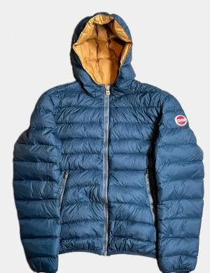 Blå pufferjacket med huva från Colmar - Snygg blå pufferjacket från Colmar med huva och kontrastfärgad insida i beige. Jackan har två sidofickor med dragkedja och ett Colmar-märke på vänster ärm. Perfekt för kalla dagar och har en modern, puffad look. Storlek Medium