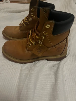 Timberland klassiska boots mocka brun - Säljer ett par ikoniska Timberland boots i brun mocka med svart vadderad kant och grov sula. Skorna har gul- och brunmönstrade snören, metallöljetter och tydlig Timberland-logga på sidan. Perfekta för dig som vill ha en tidlös och street-inspirerad look.