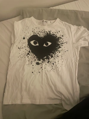 Vit Comme des Garçons Play t-shirt - Säljer en vit t-shirt från Comme des Garçons Play med det ikoniska hjärtat med ögon i svart och coolt splattermönster på framsidan. T-shirten har rund hals och korta ärmar, perfekt för dig som gillar streetstyle och grafiska prints.