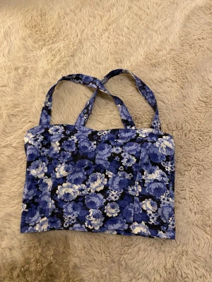 Blommig blå linnetopp  - Säljer en söt linnetopp med axelband som korsas i ryggen. Den är i strl S men passar som XS. Toppen har blommigt mönster i blått, vitt och svart. Toppen har en rak och croppad passform, perfekt för att ge din outfit en färgklick💞