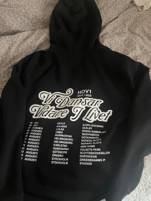 Svart Hov1 hoodie med turnétryck - Svart hoodie från Hov1 med turnétryck 'Vi Dansar Vidare I Livet' på ryggen och liten kronlogga framtill. Klassisk huva med snörning och stor magficka. Det går och pruta med priset!