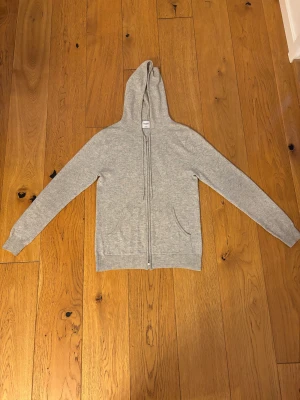 Grå cashmere hoodie från John Henric - Mjuk och lyxig ljusgrå hoodie från John Henric i 100% cashmere. Tröjan har dragkedja framtill, två fickor och huva. Perfekt för dig som vill ha en stilren och bekväm look med premiumkänsla. Hoodien är använd ett få tal gånger och köptes för 3 veckor sen. Säljer då jag har tröttnat på den 