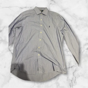 Blå rutig skjorta från Polo Ralph Lauren - Klassisk rutig skjorta från Polo Ralph Lauren med diskret rutigt mönster och broderad logga på bröstet. Skjortan har lång ärm, knappar framtill och en snygg krage. Perfekt för dig som gillar stilrena och tidlösa plagg. Custom Fit