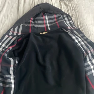 Svart teddyjacka från Burberry - Svart teddyjacka från Burberry med klassiskt rutigt foder i huvan och insidan, dragkedja framtill och broderad Burberry-logga på bröstet. Jackan har en hood och coola detaljer som silverfärgad zip. Perfekt för dig som gillar stilren design med lyxig touch.