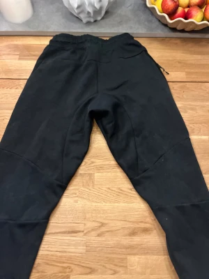 Svarta mjukisbyxor från Nike - Svarta mjukisbyxor från Nike med elastisk midja och snörning. Byxorna har en tapered passform och är tillverkade i mjukt bomullsmaterial. Nike-logga på vänster lår och diskreta detaljer längs benen. Perfekta för chill eller träning. Storlek medium men passar S är man längre än 170 kommer dom inte passa 