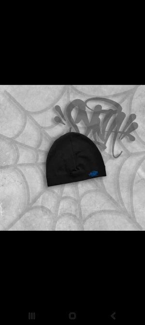 Svart mössa med blå logga (Syna world) - Svart mössa med stilren blå SYNA-logga broderad framtill. Enkel och clean design som passar till allt. Mjuk och skön passform, perfekt för kalla dagar. Materialet känns som bomull eller syntet.