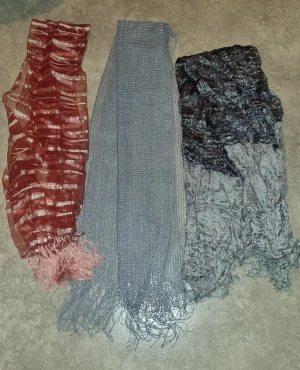 Tre snygga halsdukar i olika färger - Säljer tre halsdukar: en röd och beige med randigt mönster och fransar, en grå stickad med långa fransar och en svart/grå med blommigt mönster och skrynklig struktur. Perfekt för dig som vill variera din stil och gillar unika accessoarer.