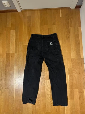 Svarta cargopants från Carhartt - Svarta cargopants från Carhartt Work In Progress med avslappnad passform. Byxorna har flera fickor, inklusive bakfickor med Carhartt-logga, och är tillverkade i slitstark bomull. Perfekt för dig som gillar streetwear och funktionella detaljer. (1299kr nypris)