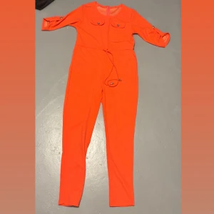 Orange/rosa jumpsuit köpt utomlands  - Jumpsuiten har en dragkedja i ryggen och ett knytband i midjan för extra passform, även knapp detaljer. Materialet känns mjukt och har en lätt stretch. (Den ser mer rosa/orange ut i verkligheten än bara orange) 