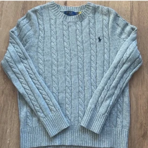 Grå kabelstickad tröja Polo Ralph Lauren - Kabelstickad grå tröja från Polo Ralph Lauren med rund halsringning och broderad logga på bröstet. Tröjan har långa ärmar och breda ribbade muddar vid ärmslut och nederkant. Perfekt för lager-på-lager under kyliga dagar.