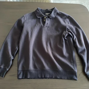 Svart pikétröja från H&M - Klassisk svart pikétröja från H&M med krage och tre knappar framtill. Tröjan har lång ärm och är tillverkad i mjuk bomull, perfekt för en clean och enkel stil. Regular fit som sitter snyggt utan att vara för tight.