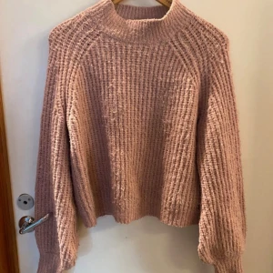 Rosa stickad tröja Pernilla Wahlgren - Mysig rosa stickad tröja från Pernilla Wahlgren med grov ribbad struktur och hög hals. Tröjan har långa ärmar och en liten metallbricka med ananasdetalj nertill. Perfekt för kyliga dagar och enkel att styla till jeans eller kjol. Den är i storlek L men passar bättre åå S/M. Väldigt nopprig