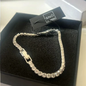 Silverfärgat armband SNÖ of Sweden - Stilrent armband från SNÖ of Sweden i silverfärg med gnistrande stenar längs hela bandet. Klassisk tennisarmbandsdesign med fyrkantiga länkar och smidig låsdetalj. Perfekt för dig som gillar glittriga accessoarer med lyxig känsla.