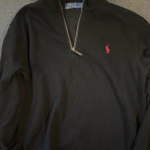 Svart half zip tröja från Polo Ralph Lauren - Svart långärmad tröja från Polo Ralph Lauren med half zip och röd broderad logga på bröstet. Klassisk och stilren modell i bomull, perfekt för lager-på-lager och chill vardagsstil.